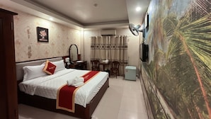 Habitación de hotel con cama, escritorio, silla, mesita, espejo, televisor montado en la pared y un mural tropical en la pared.