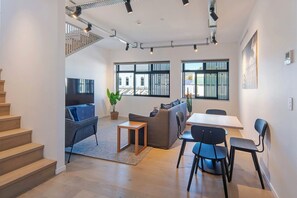 Una sala de estar moderna con una escalera, un televisor de pantalla plana, un sofá gris, una mesa de comedor con sillas y ventanas grandes con persianas.