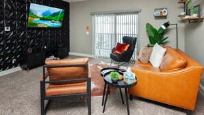 Un salon moderne avec un canapé en cuir, un fauteuil noir, un téléviseur fixé sur un mur orné d’un motif géométrique et une grande fenêtre avec des stores.