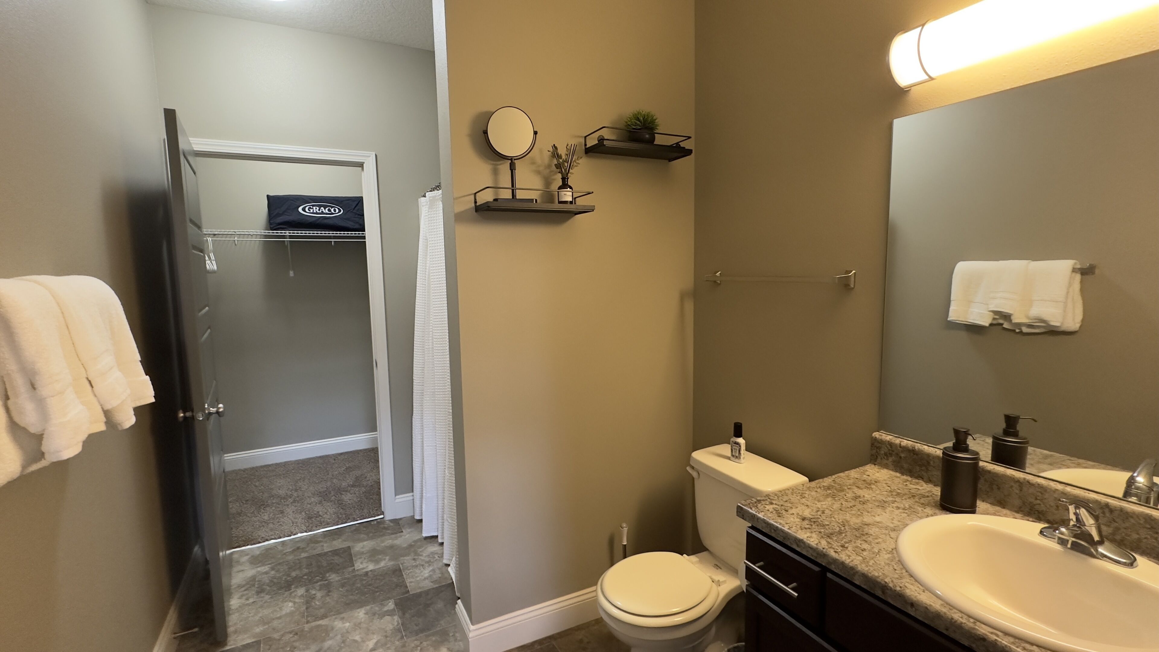 A bathroom with a toilet, sink, and shower. There is a towel rack with a towel, a mirror, and a shelf with a plant.