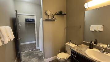 A bathroom with a toilet, sink, and shower. There is a towel rack with a towel, a mirror, and a shelf with a plant.