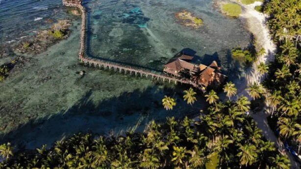 An aerial view of a coastal property with a pier leading to a building, surrounded by palm trees and clear blue waters. - Malayah Hostel (General Luna)