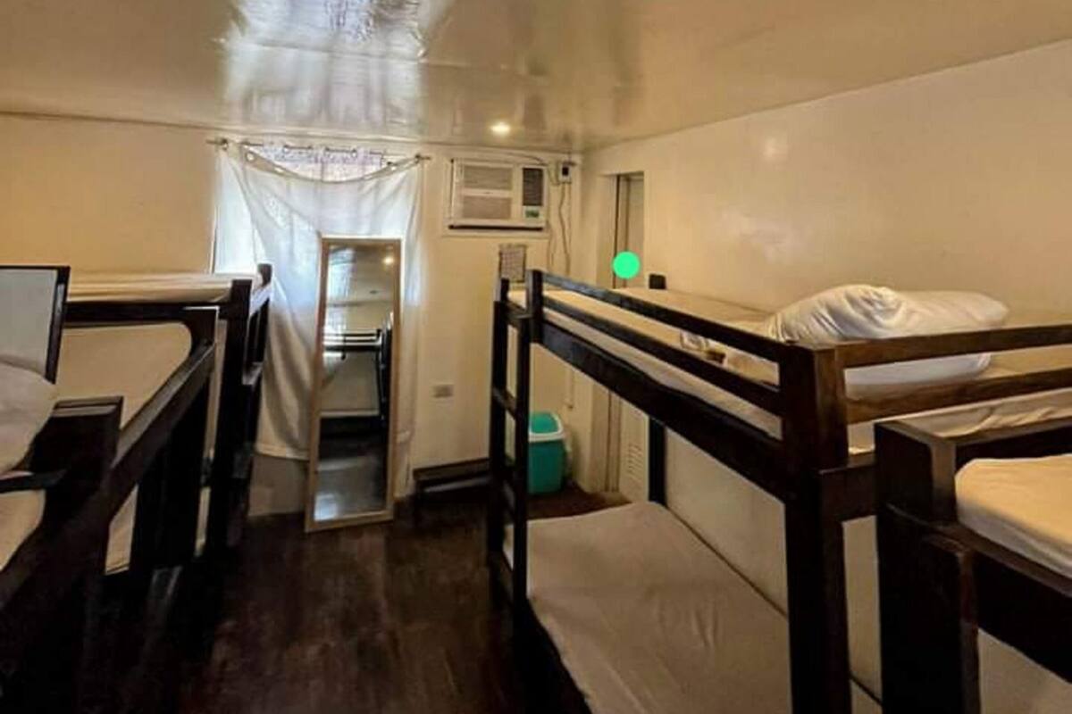 A narrow, dimly lit room with bunk beds, a small kitchenette, and a curtained window.