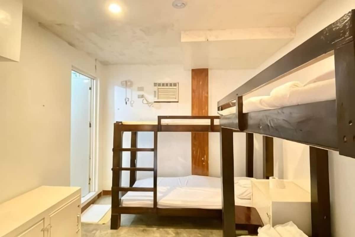 A bunk bed room with a wooden bunk bed, a small bedside table, a white dresser, and a door leading to another room.