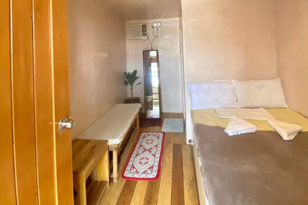 A small, compact room with a single bed, a wooden bench, a small table, and a red patterned rug.
