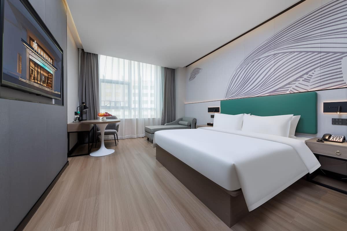 A modern hotel room with a large bed, a desk, a chair, and a view of the city through large windows.