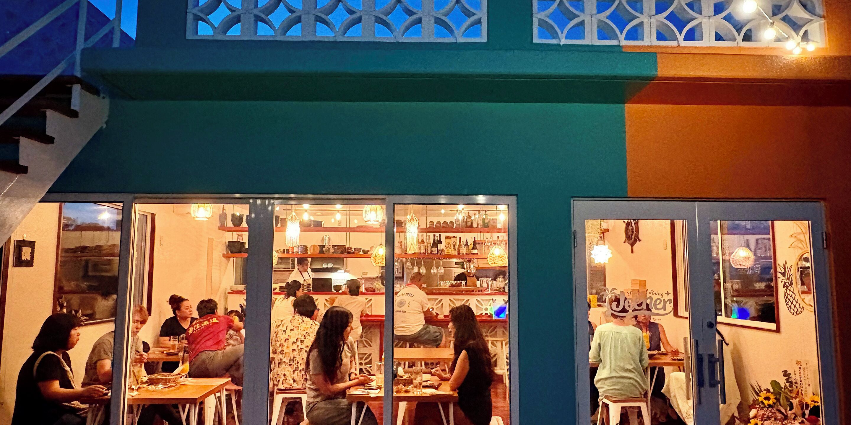 An outdoor dining area with tables and chairs, a counter with a person working behind it, and a decorative pineapple sign on the wall.