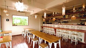 Zona de bar con mesas de madera y taburetes blancos, un ventanal con vistas a la vegetación y lámparas colgantes.