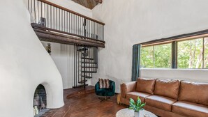 Ein Wohnzimmer mit einem braunen Ledersofa, einem runden Holztisch und einem grünen Sessel. Ein Kamin aus Ziegelsteinen ist sichtbar, und eine Treppe mit einem Hollauf führt in eine obere Ebene.