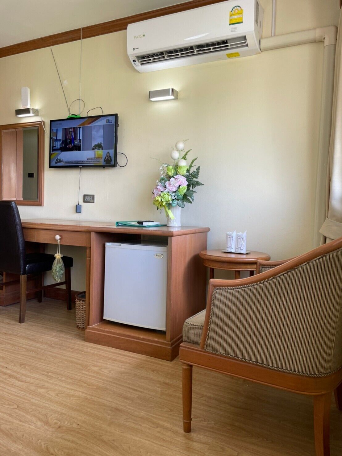 A compact room with a wooden desk, a small refrigerator, a television mounted on the wall, a chair, and a decorative vase with flowers.