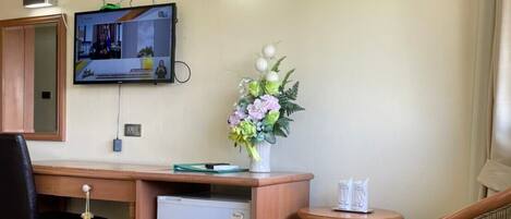 A compact room with a wooden desk, a small refrigerator, a television mounted on the wall, a chair, and a decorative vase with flowers.