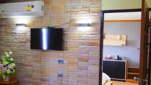 Een kamer met een wand in steenmotief, een wandgemonteerde flatscreen-tv, een houten nachtkastje met een bloevas, een bed en een klein houten kastje.