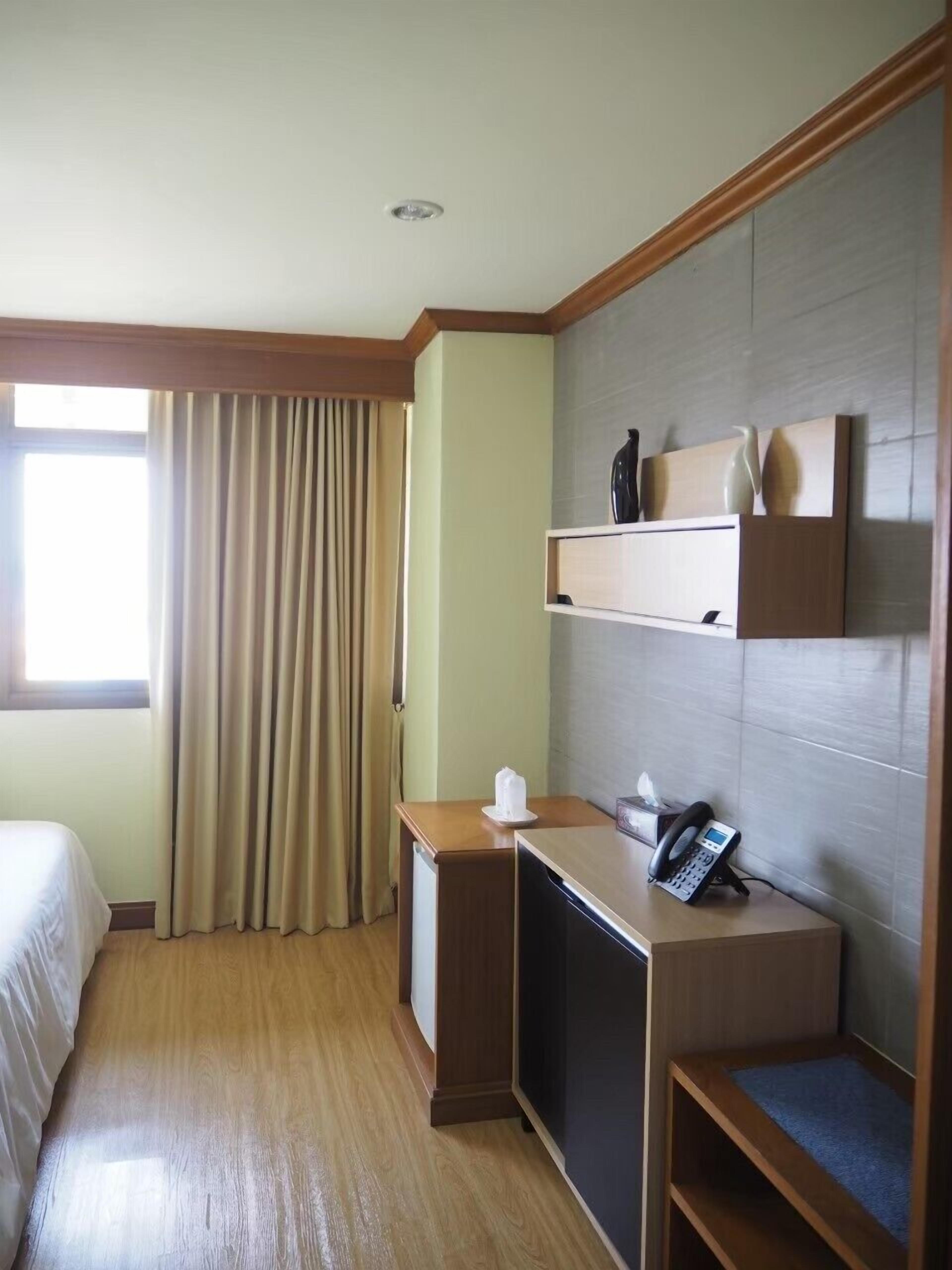 A hotel room with a bed, a desk with a phone, a small shelf with decorative items, and a window with curtains.