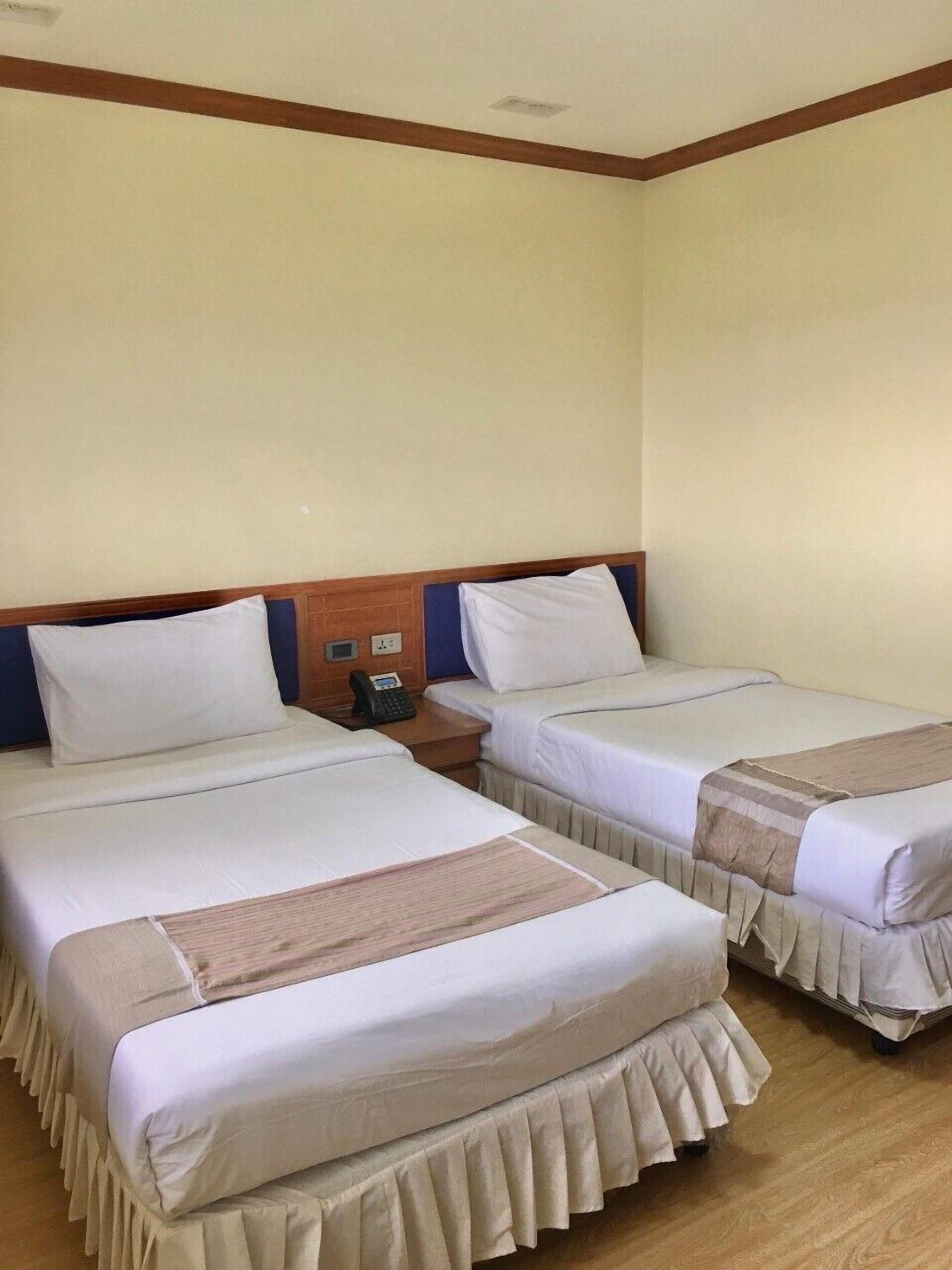 A hotel room with two single beds, wooden headboards, a small bedside table with a phone, and a plain cream-colored wall.