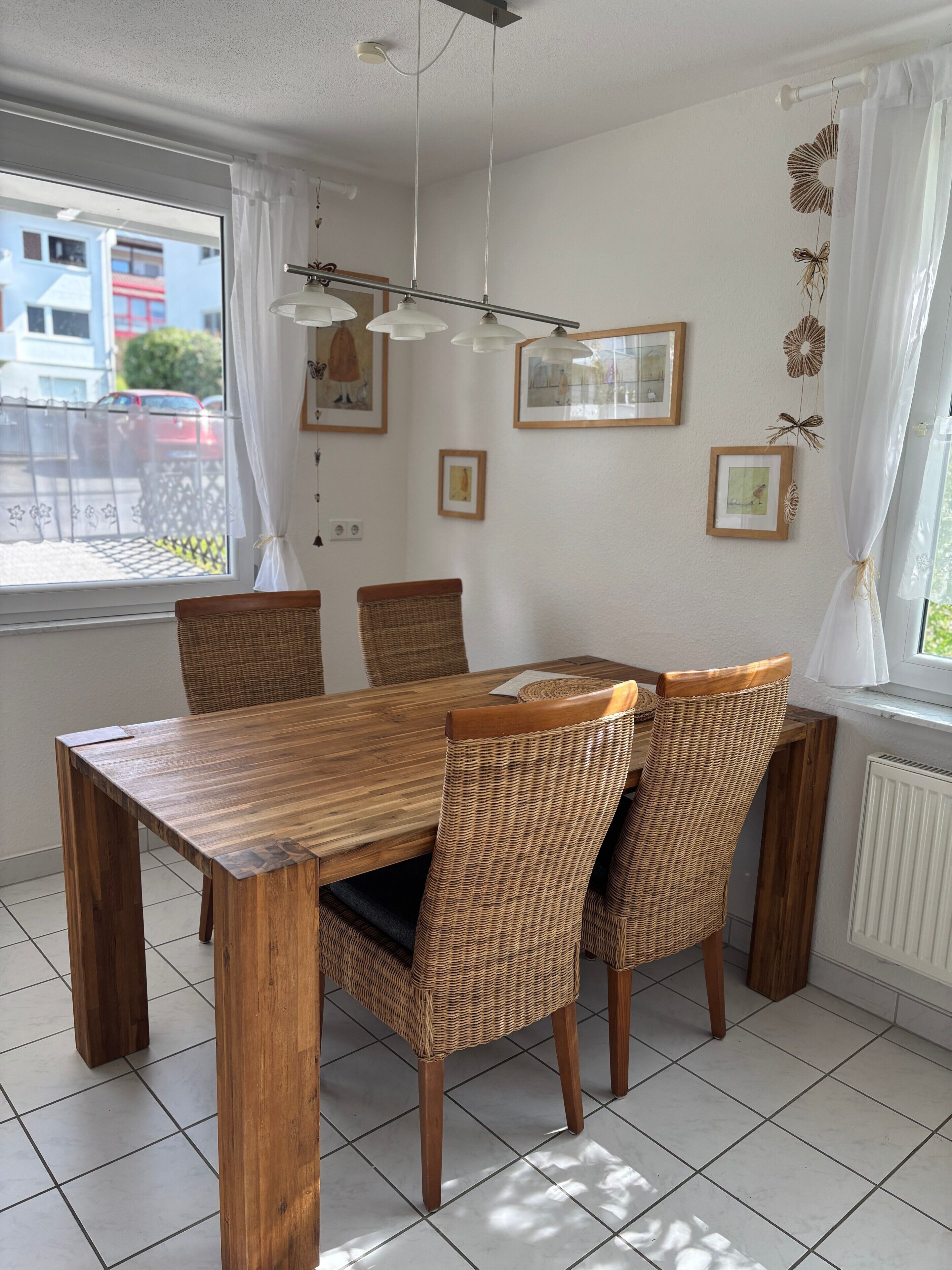 A dining area with a wooden table and wicker chairs, white curtains, and framed pictures on the wall.