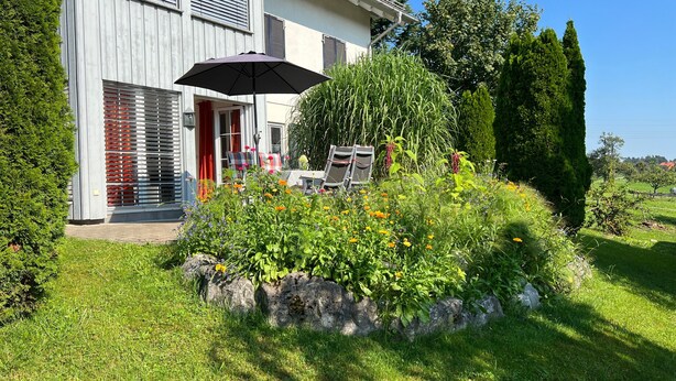 Un bâtiment moderne doté d’un espace jardin agrémenté de diverses plantes et fleurs, de mobilier d’extérieur et d’une porte rouge.