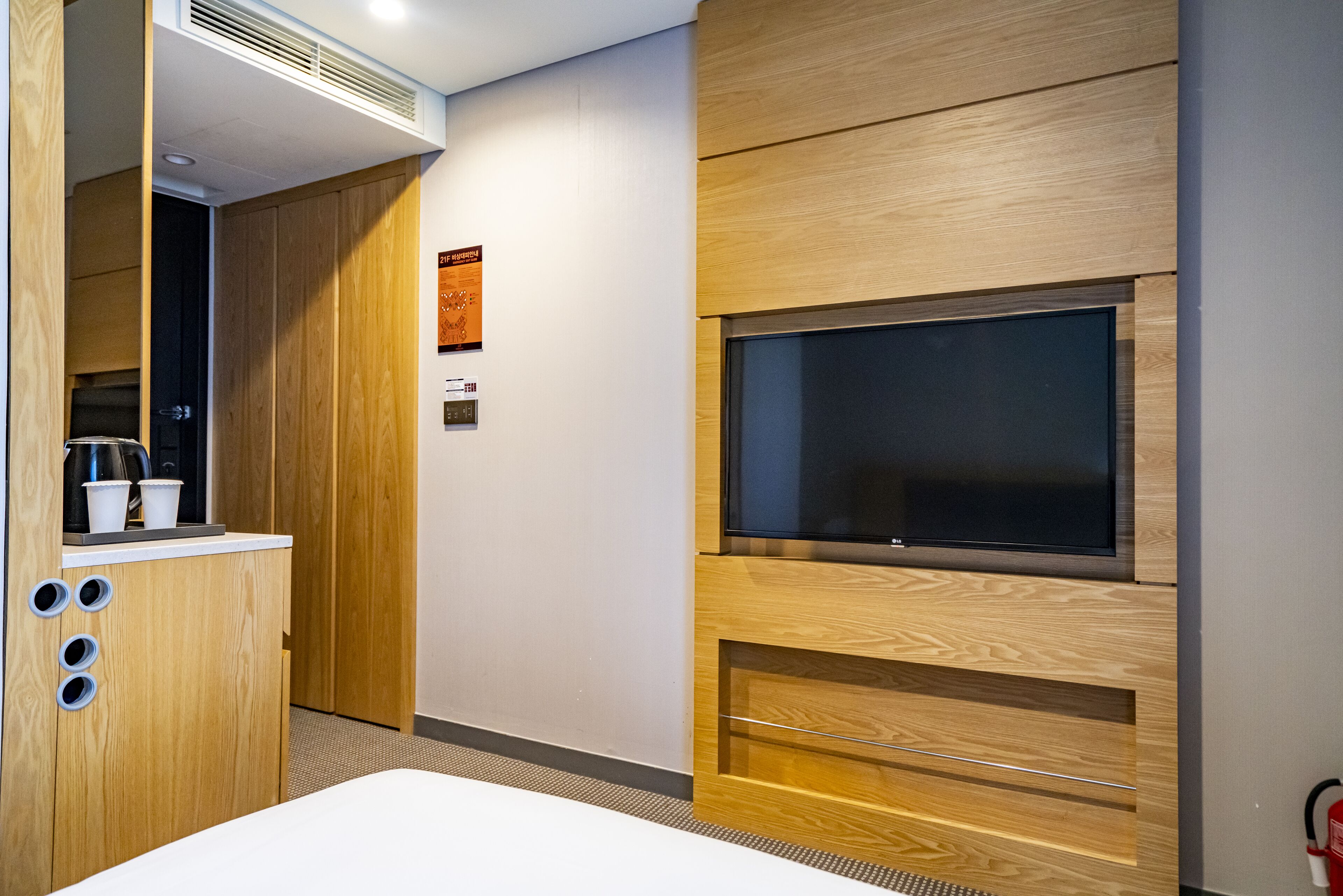 A modern hotel room with a wooden cabinet, a flat-screen TV mounted on the wall, and a small kitchenette area.