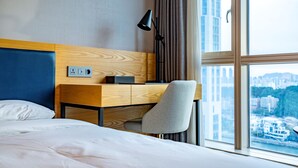 A hotel room with a bed, a desk, a chair, a lamp, and a window with a city view. - Centum Business Hotel (Busan)