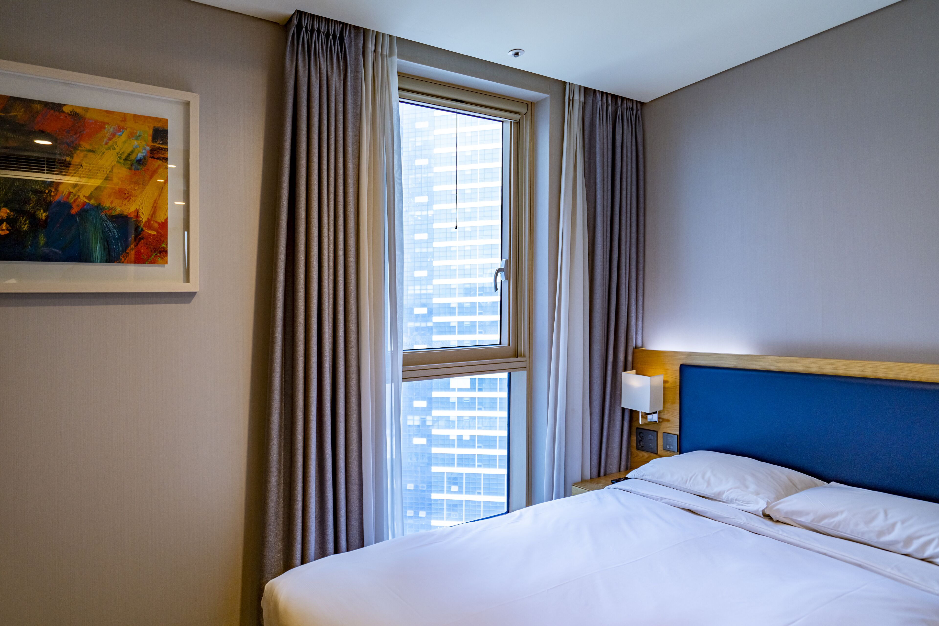 Habitación de hotel con cama, ventana con cortinas, un cuadro colgado en la pared y una mesita de noche con lámpara.