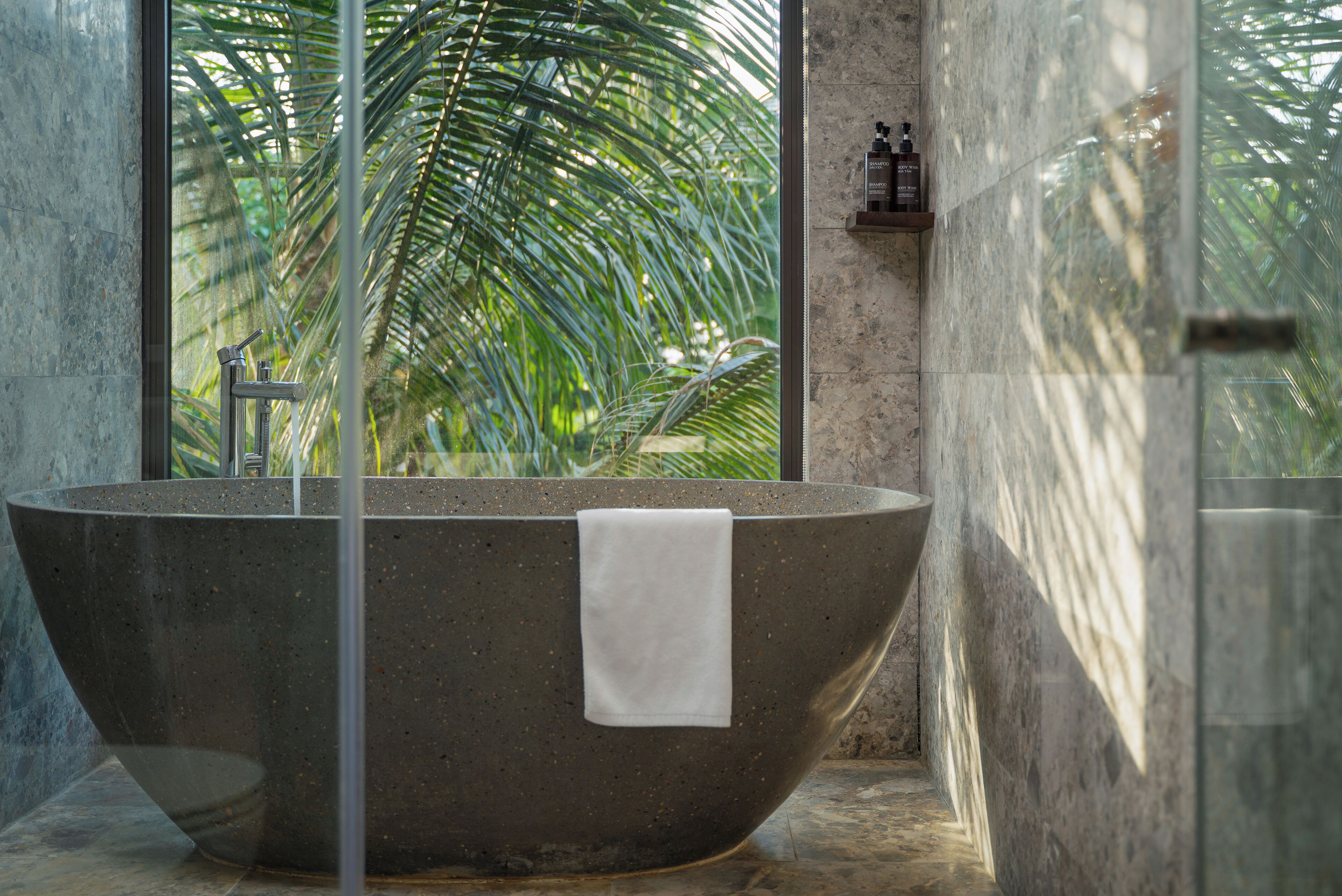 A large freestanding bathtub in a modern bathroom with a glass enclosure, a white towel hanging on the edge, and a view of palm trees through a large window.