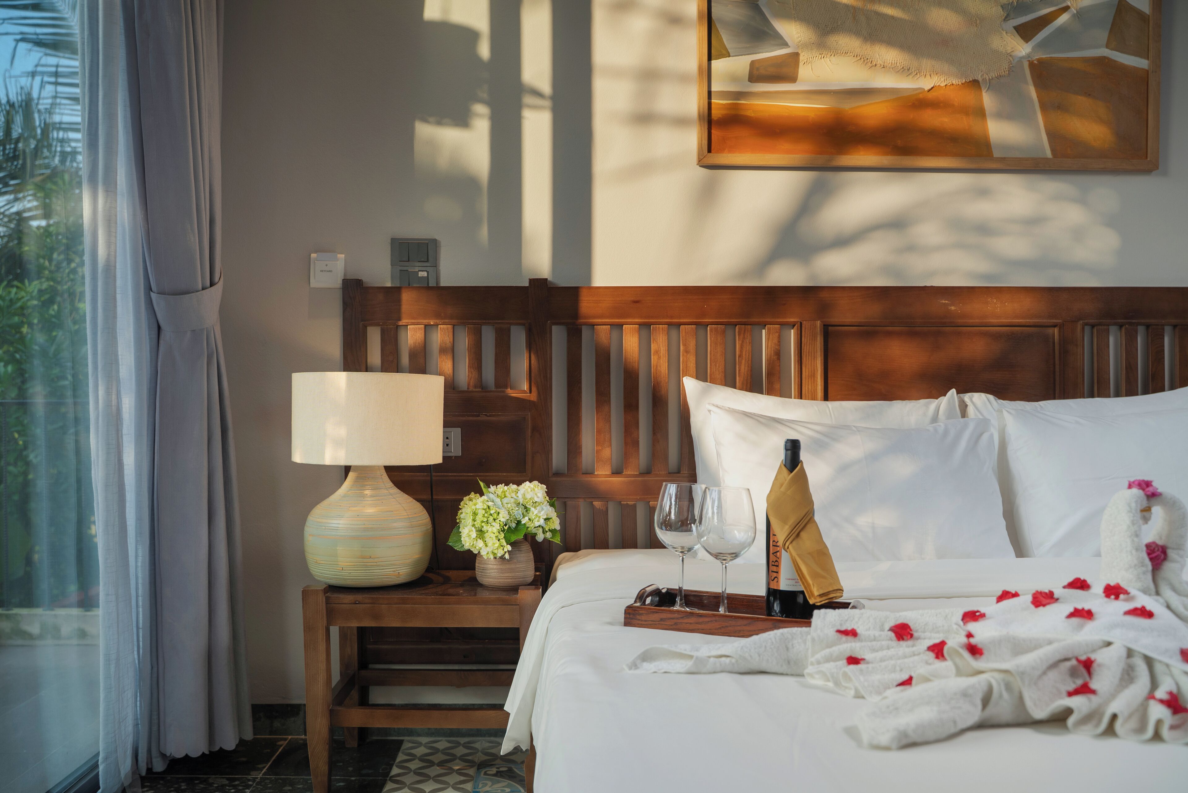 A hotel room with a wooden headboard, a bedside table with a lamp, a vase of flowers, a tray with a bottle and glasses, and a bed with white linens and rose petals.