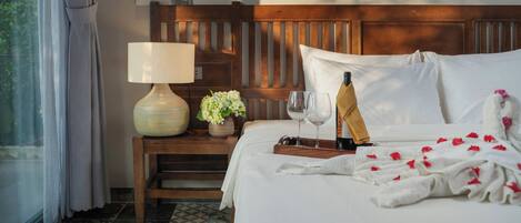 A hotel room with a wooden headboard, a bedside table with a lamp, a vase of flowers, a tray with a bottle and glasses, and a bed with white linens and rose petals.