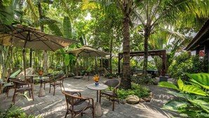 Un patio al aire libre con mobiliario de mimbre, sombrillas y suelo de piedra, rodeado de abundante vegetación y palmeras.