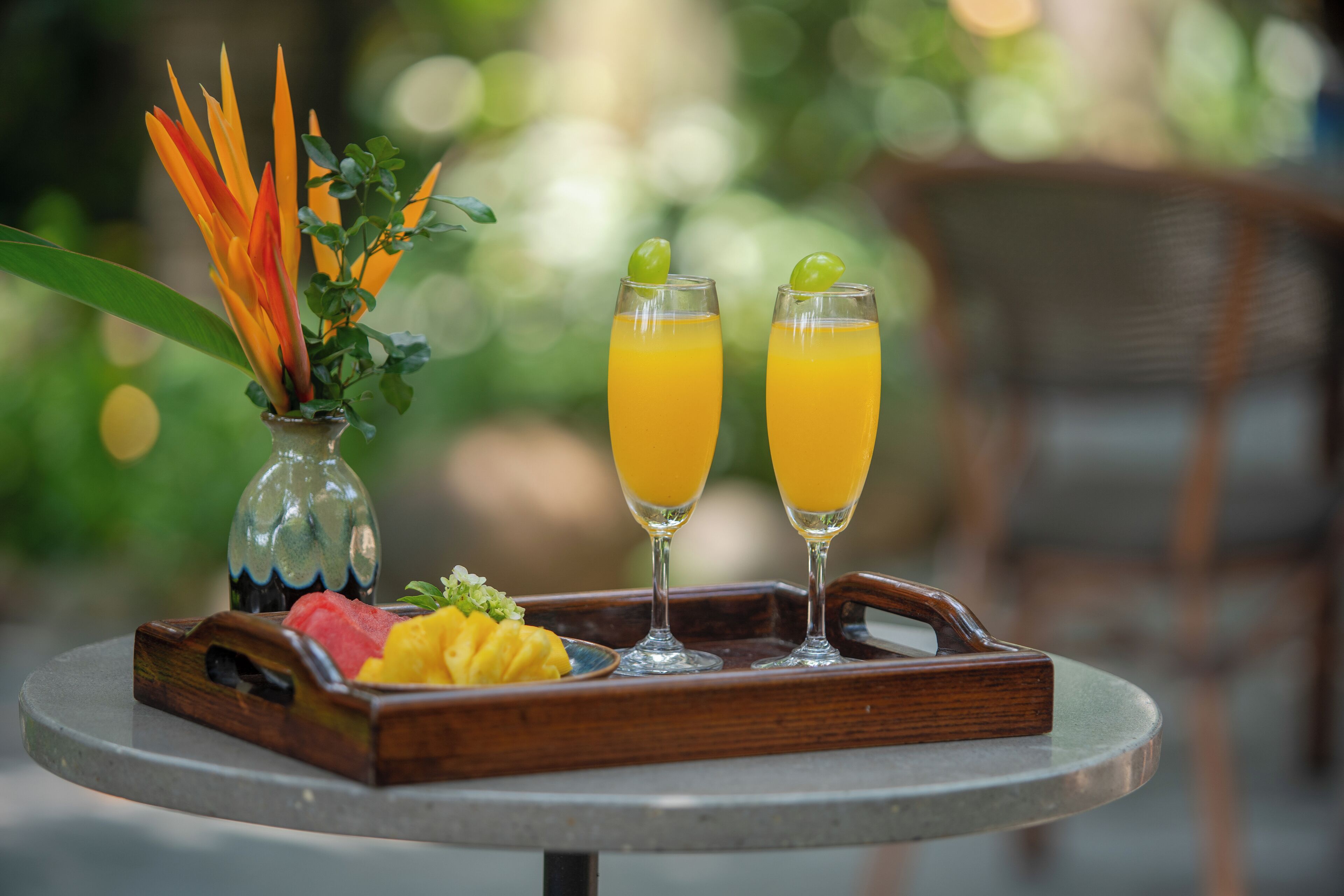 A tray with two glasses of orange juice, a small vase with orange flowers, and a wooden tray with fruit.