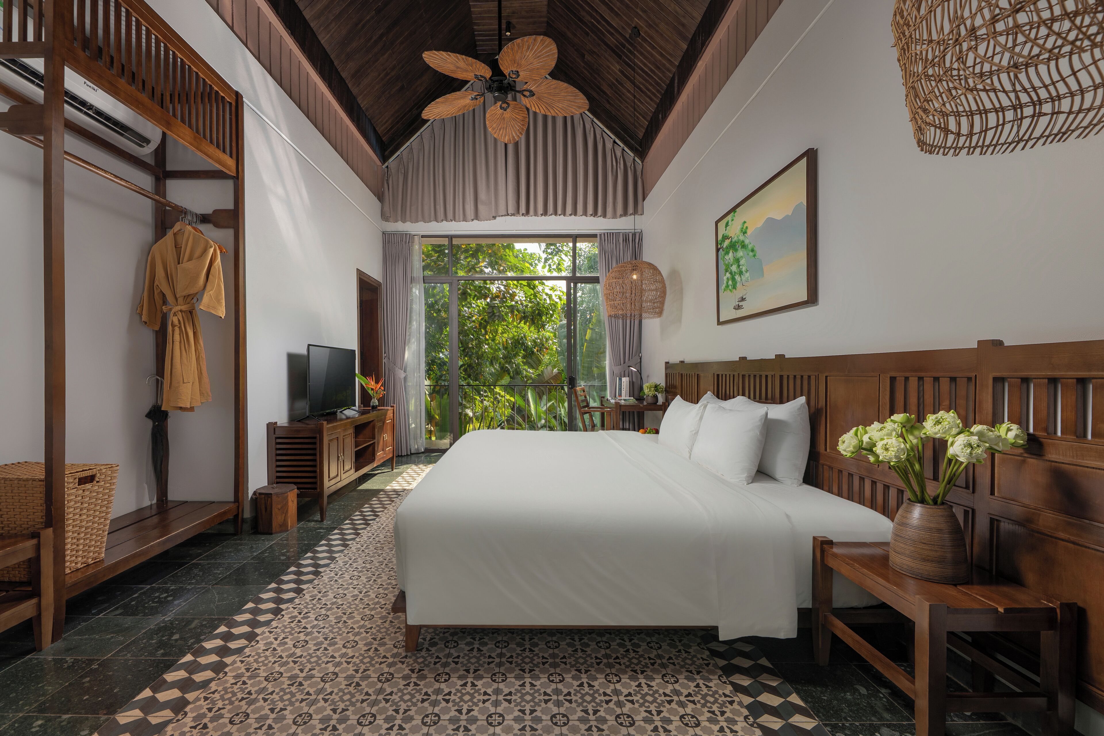 A spacious bedroom with a large bed, wooden headboard, a ceiling fan, a television, and a view of greenery outside.