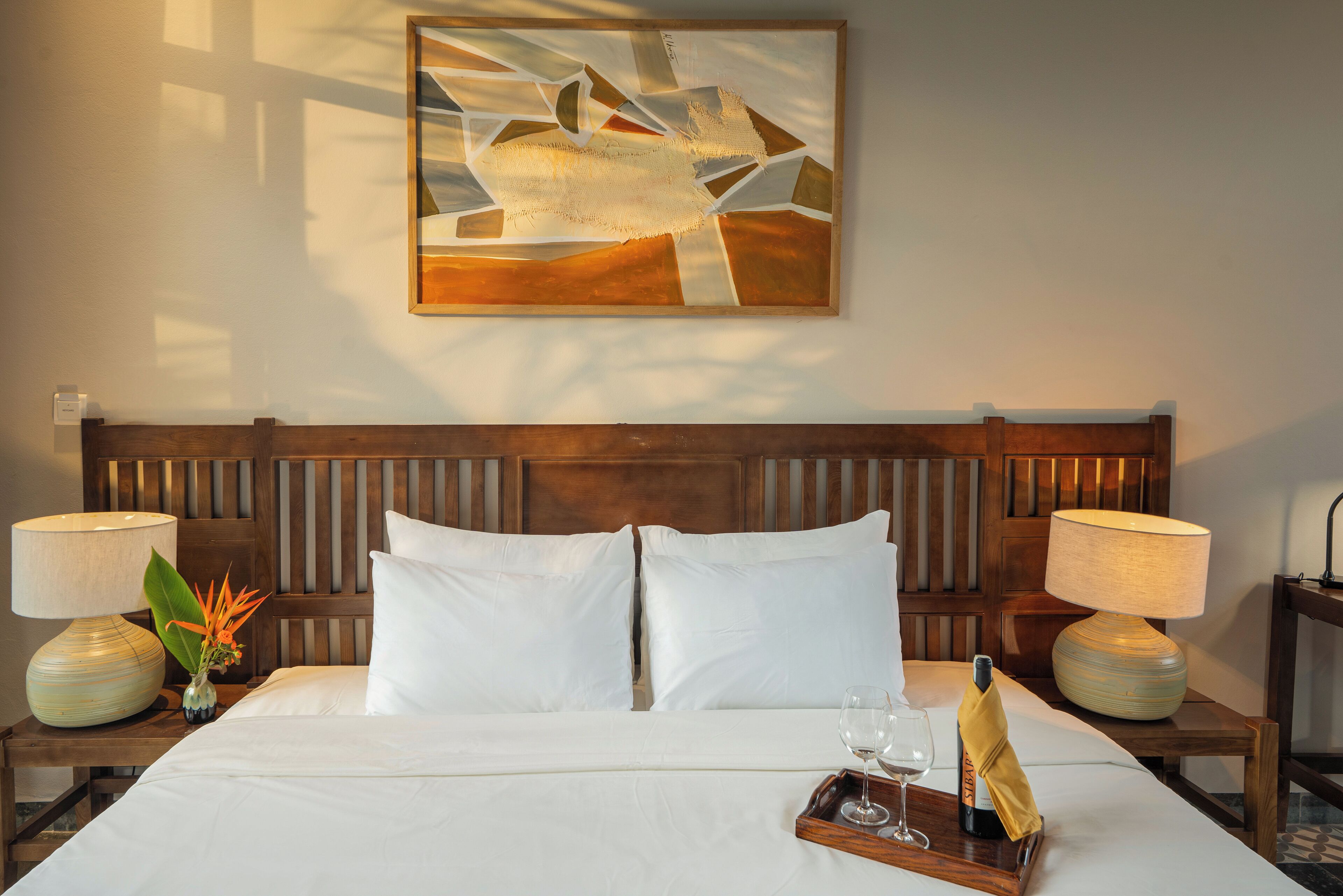 A hotel room with a wooden headboard, white bedding, two bedside lamps, a vase with flowers, and a tray with a wine glass and bottle.