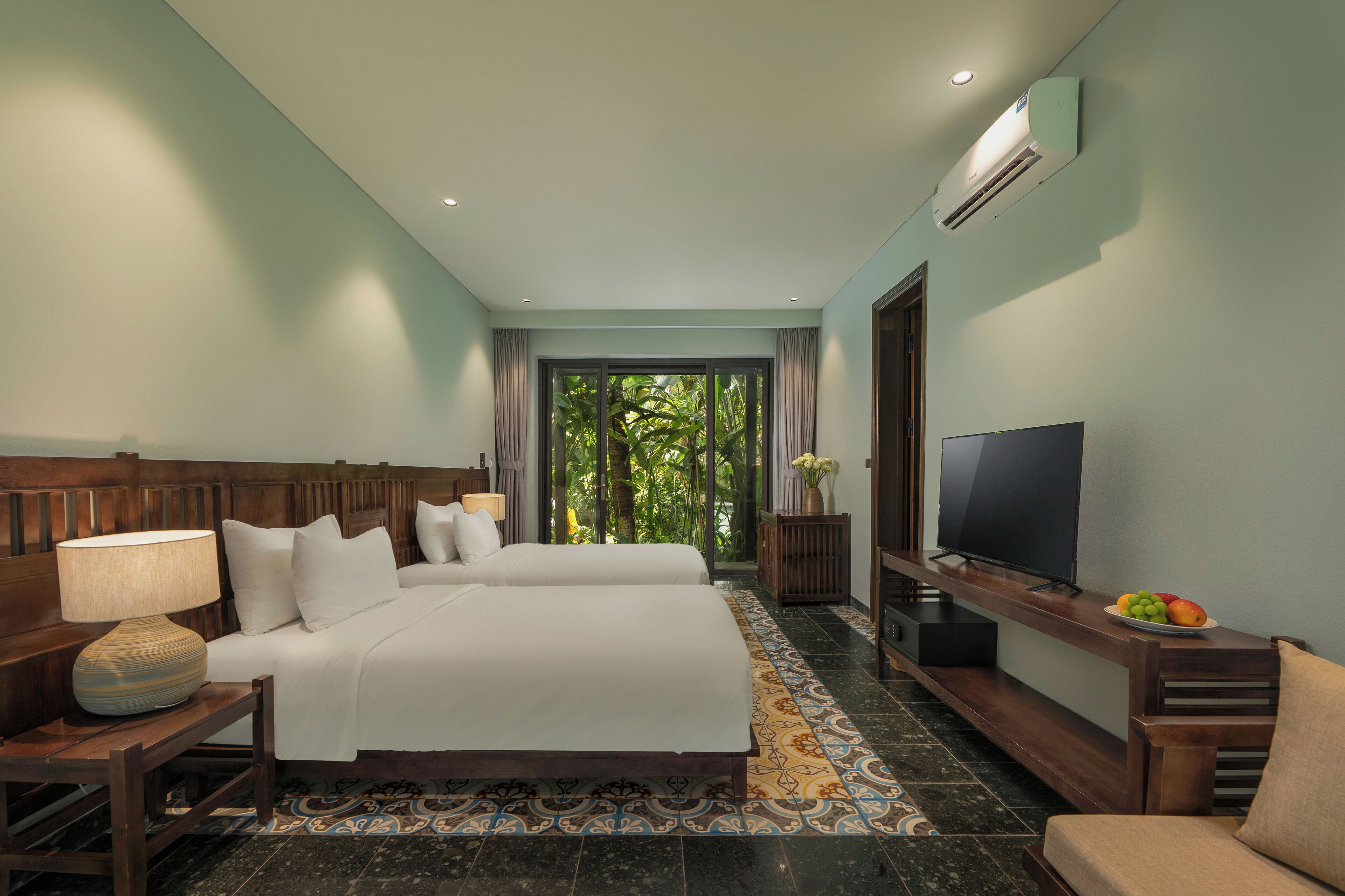 A hotel room with a large bed, a TV, a desk with a lamp, and a view of greenery through the window.