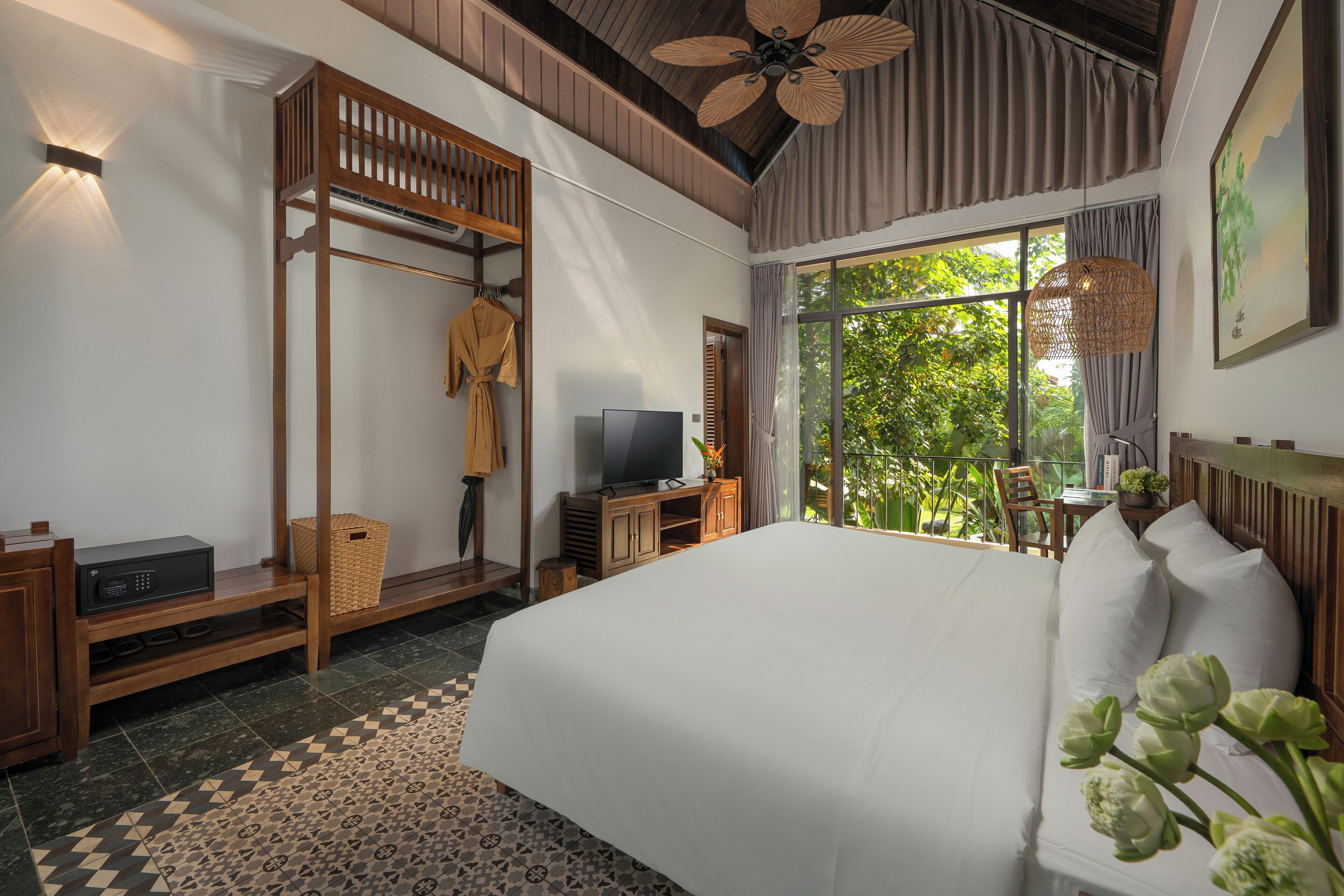 A bedroom with a large bed, a television, a wooden wardrobe, and a window with a view of greenery.