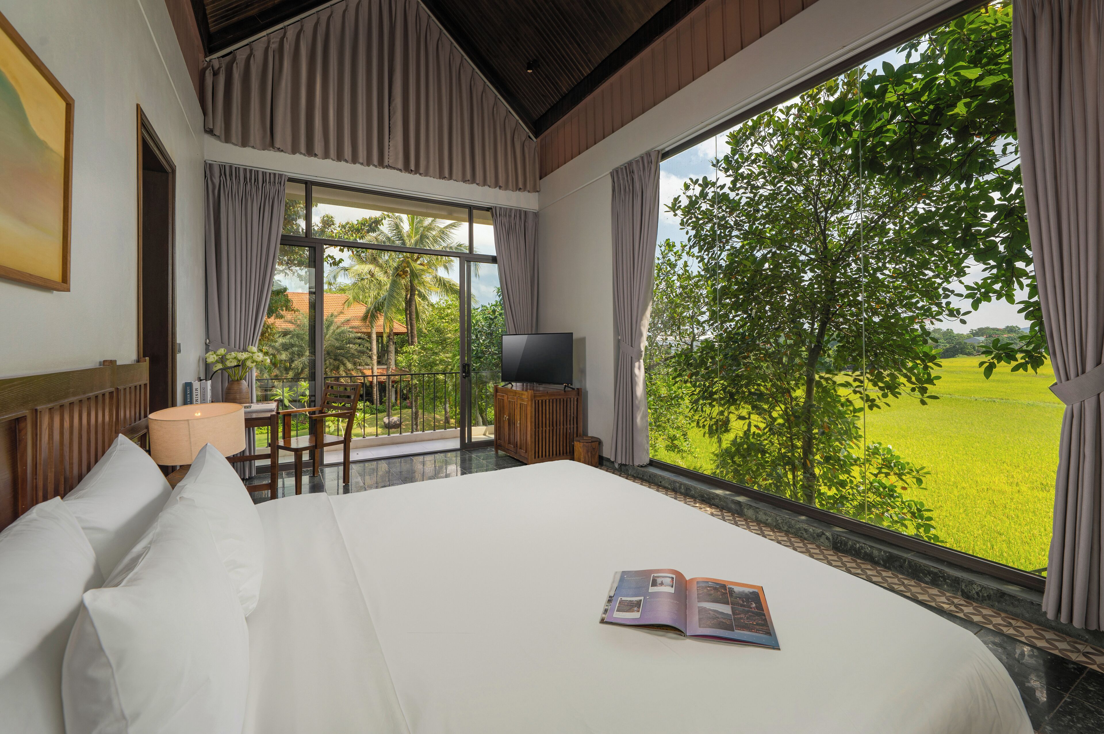 A hotel room with a large bed, a TV, a table with chairs, and a view of a green landscape.