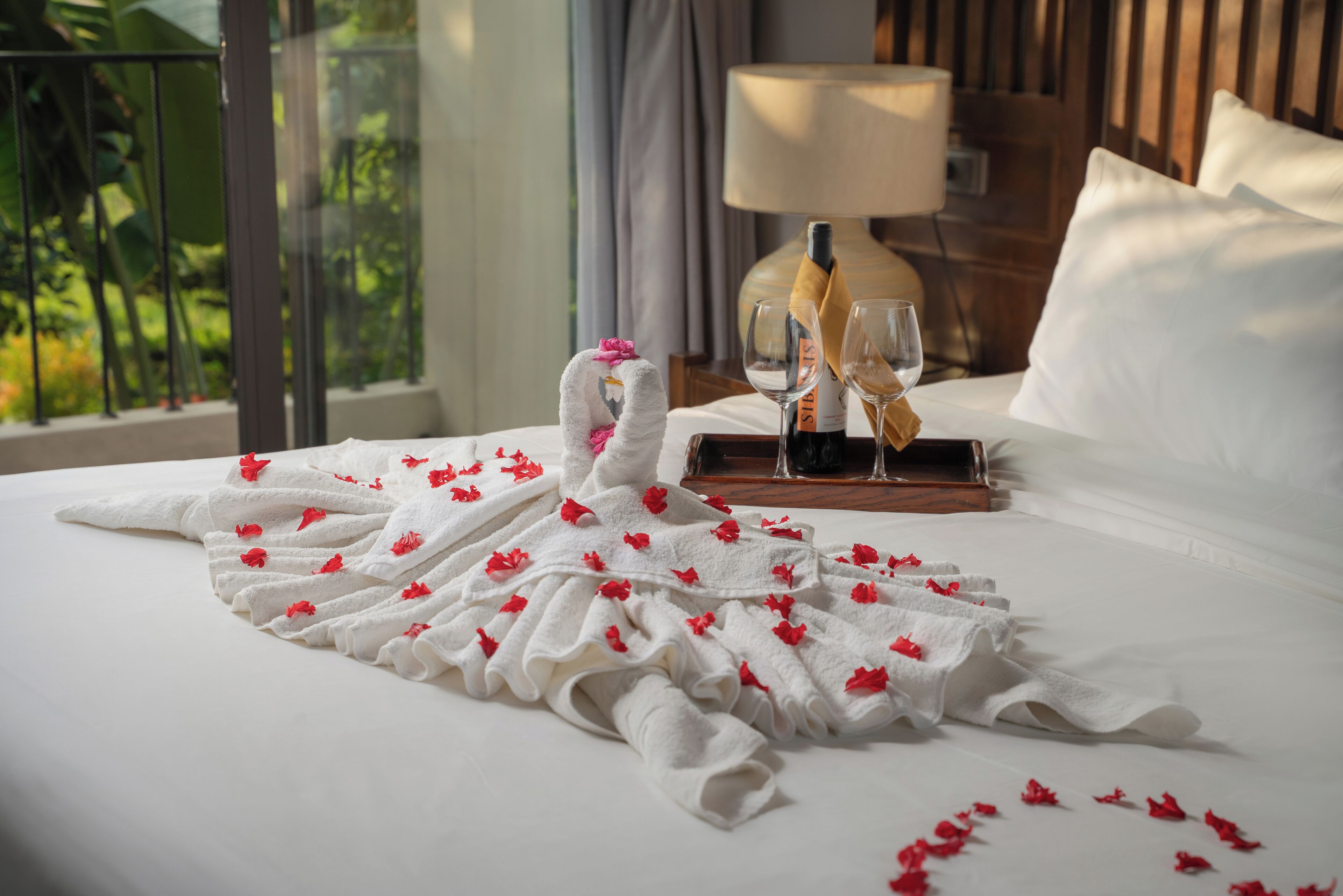 A neatly made bed with a towel arranged in the shape of a swan, a bottle of wine, and two glasses on a tray.