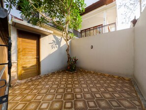 Un patio con suelo de baldosas, una puerta de madera, un árbol y un muro con un balcón.