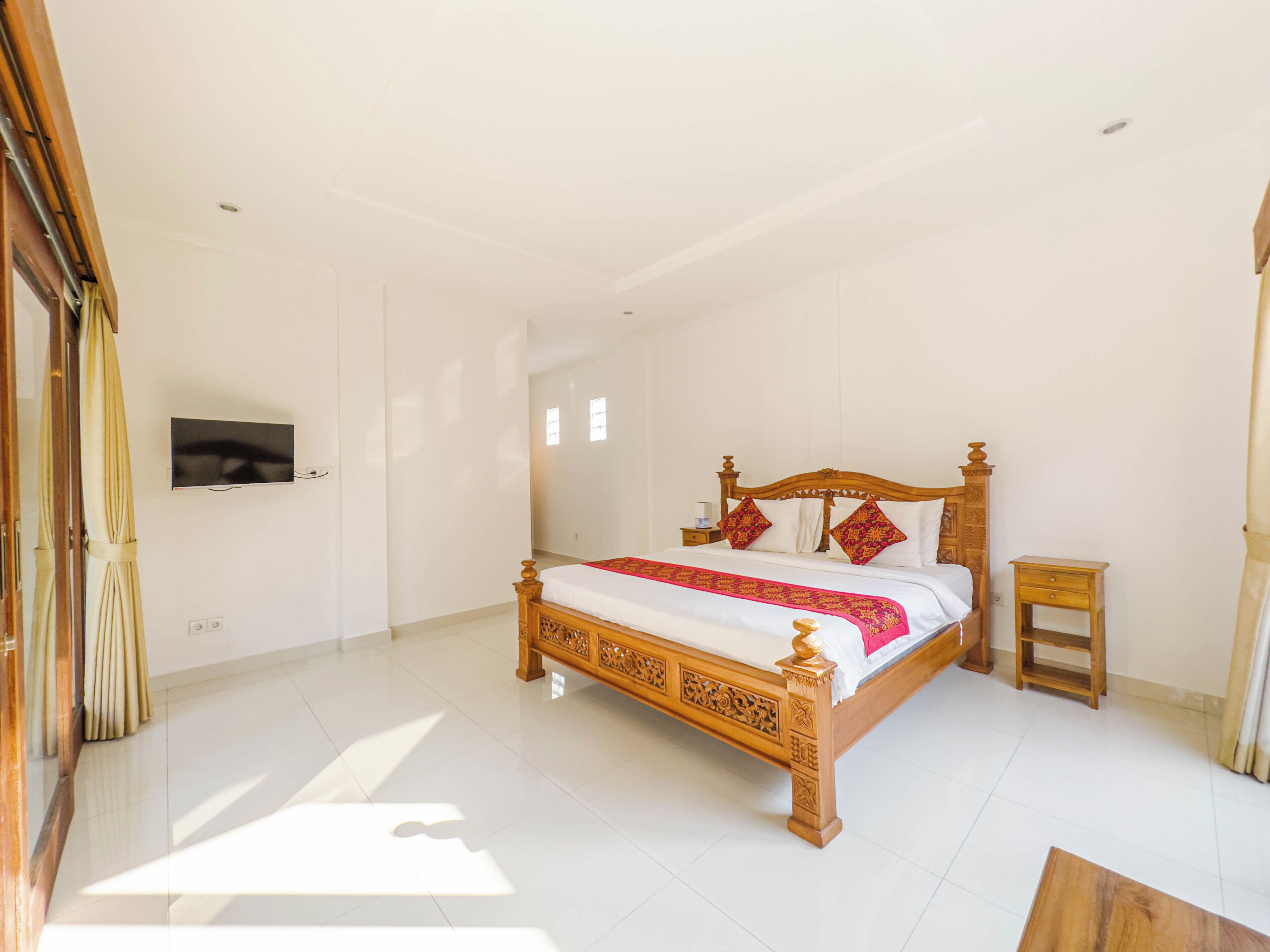 A spacious bedroom with a large bed, a wooden headboard, a small bedside table, a television mounted on the wall, and a sliding door.
