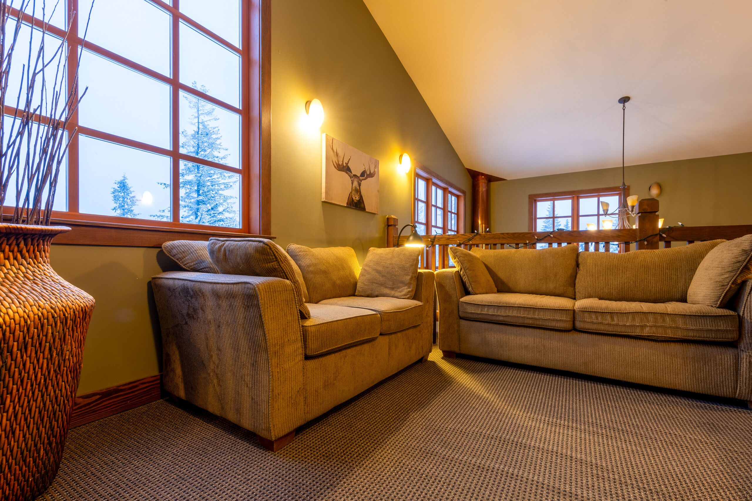 A cozy living room with two sofas, a large window with a view of trees, and a wall-mounted deer painting.