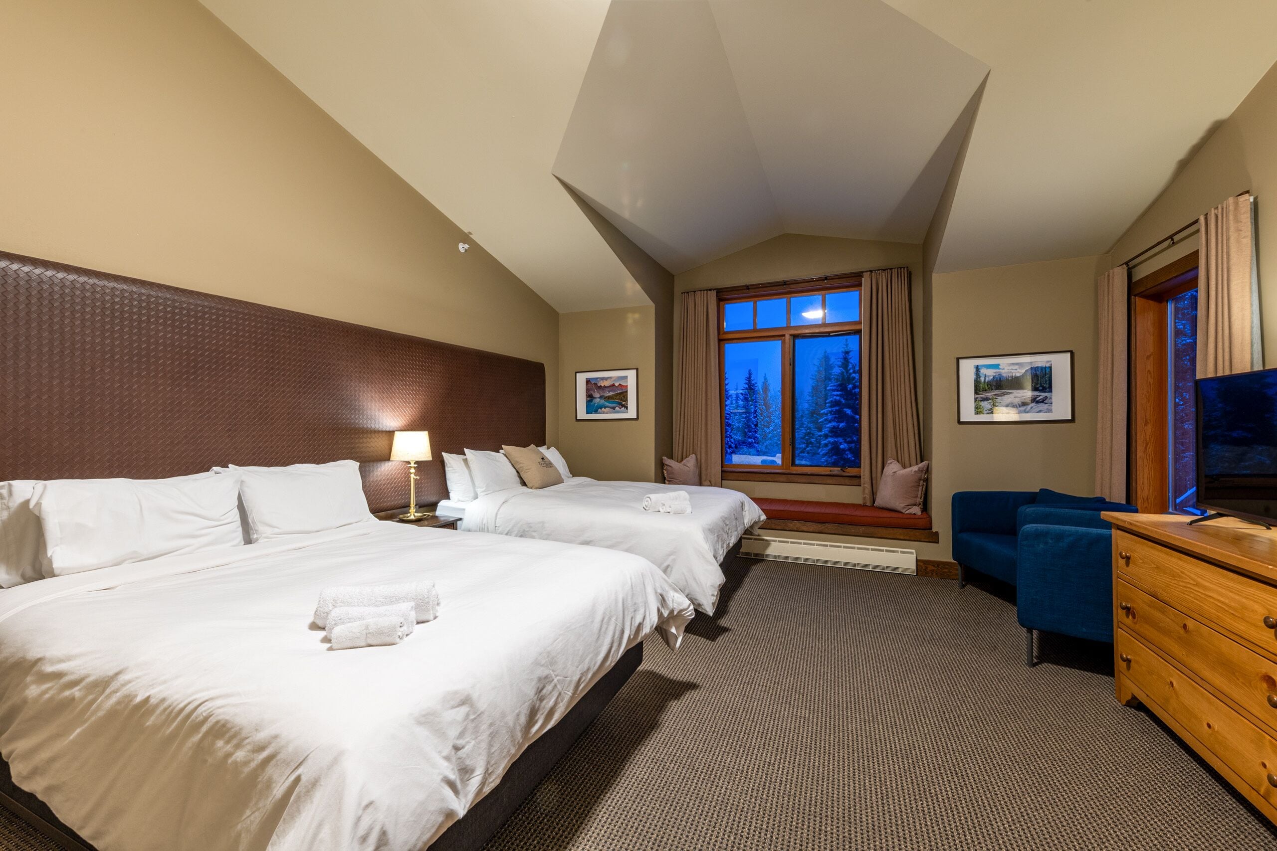 A hotel room with two beds, a TV, a dresser, and a window with a view of trees.