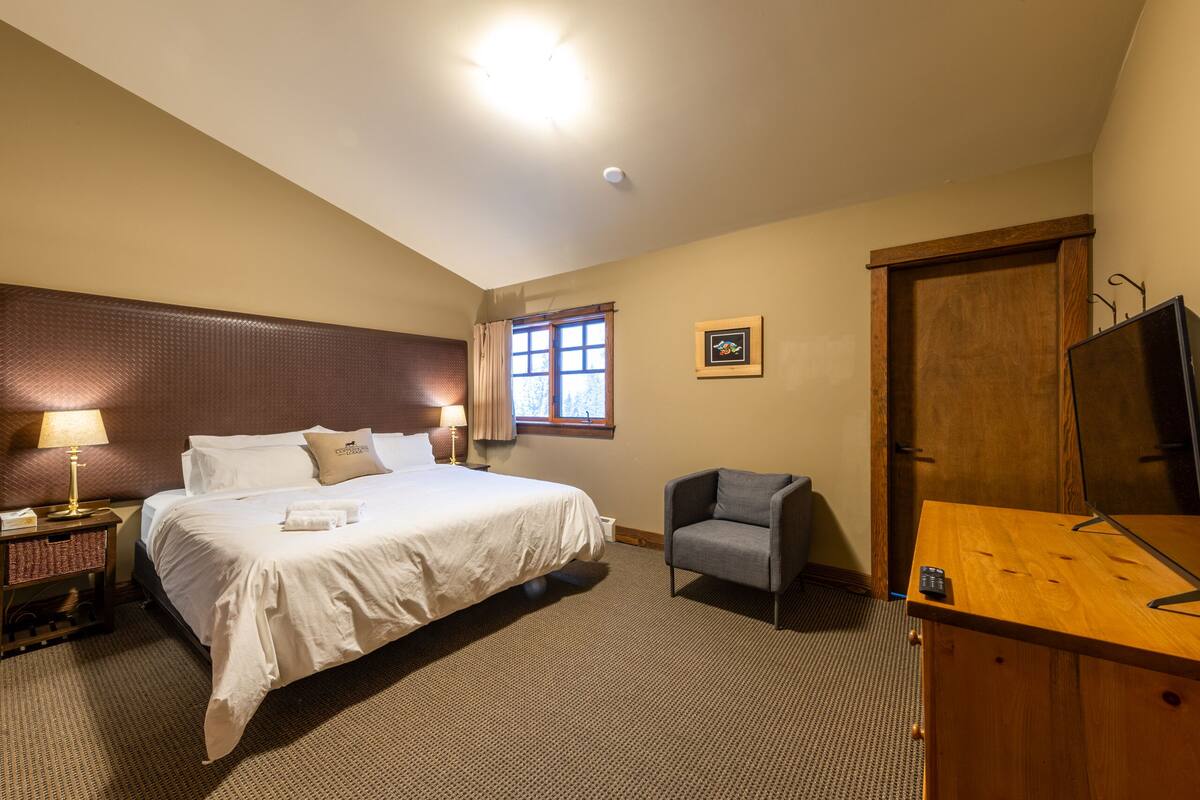 Quarto de hotel com uma cama grande, uma cadeira, uma mesa, uma televisão, uma janela e uma porta.