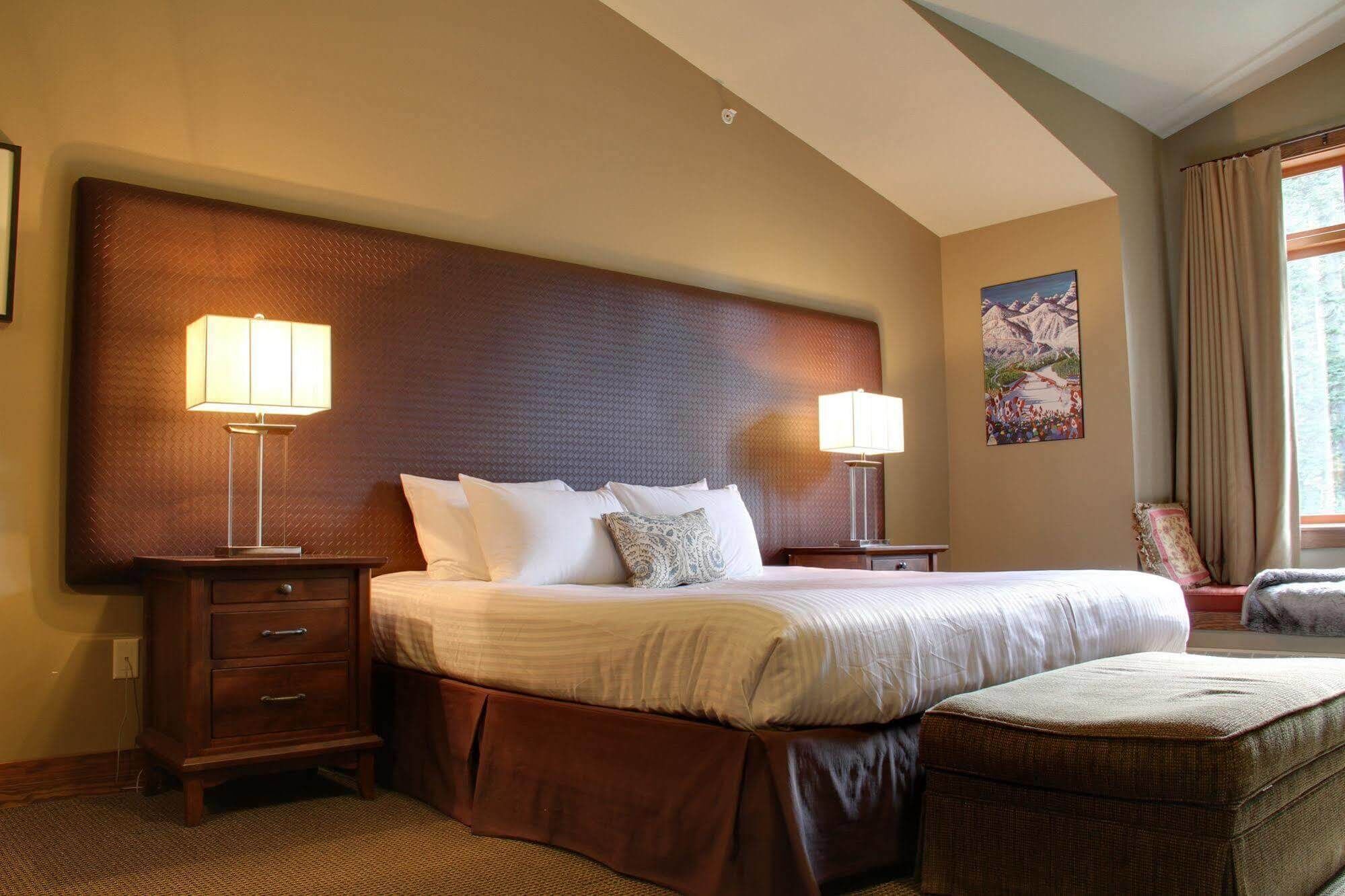 A hotel room with a large bed, two bedside lamps, a wooden headboard, a small side table, a chair, and a painting on the wall.
