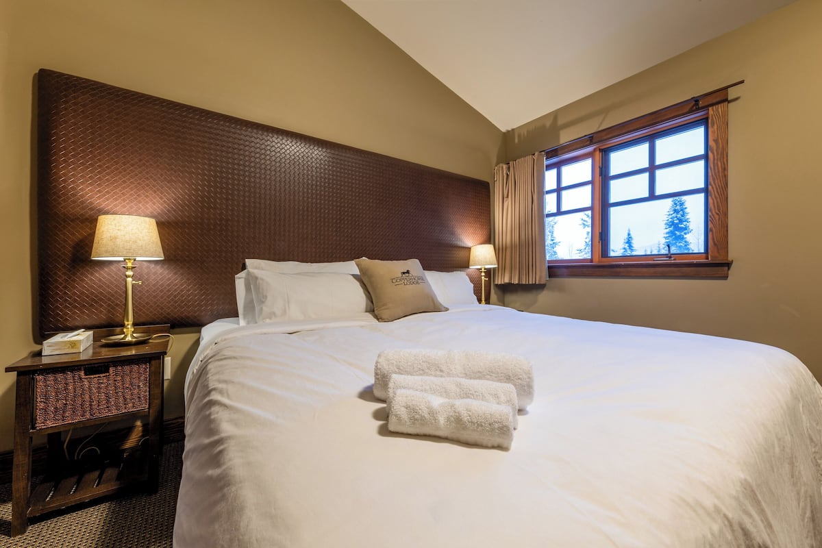 Cama bem arrumada com lençóis brancos e duas toalhas enroladas, cabeceira de madeira, mesa de cabeceira com luminária e janela com cortinas.