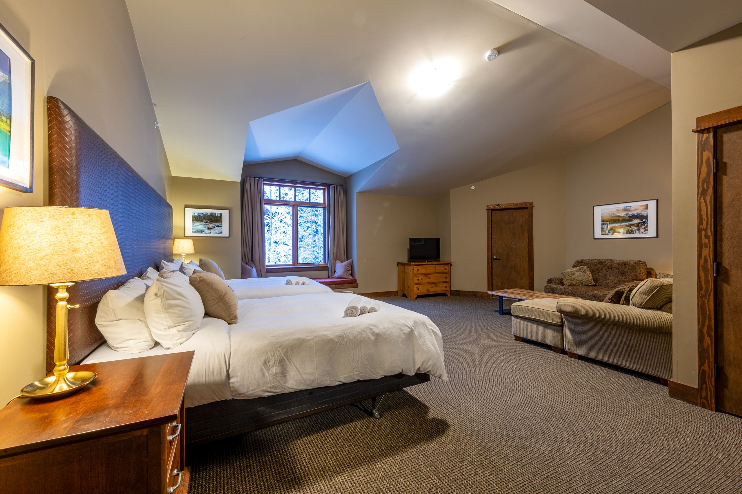 Ein Hotelzimmer mit einem großen Bett, einer Sitzecke, einem Schreibtisch mit Lampe, einem Fernseher und einem Fenster mit Blick auf Bäume.