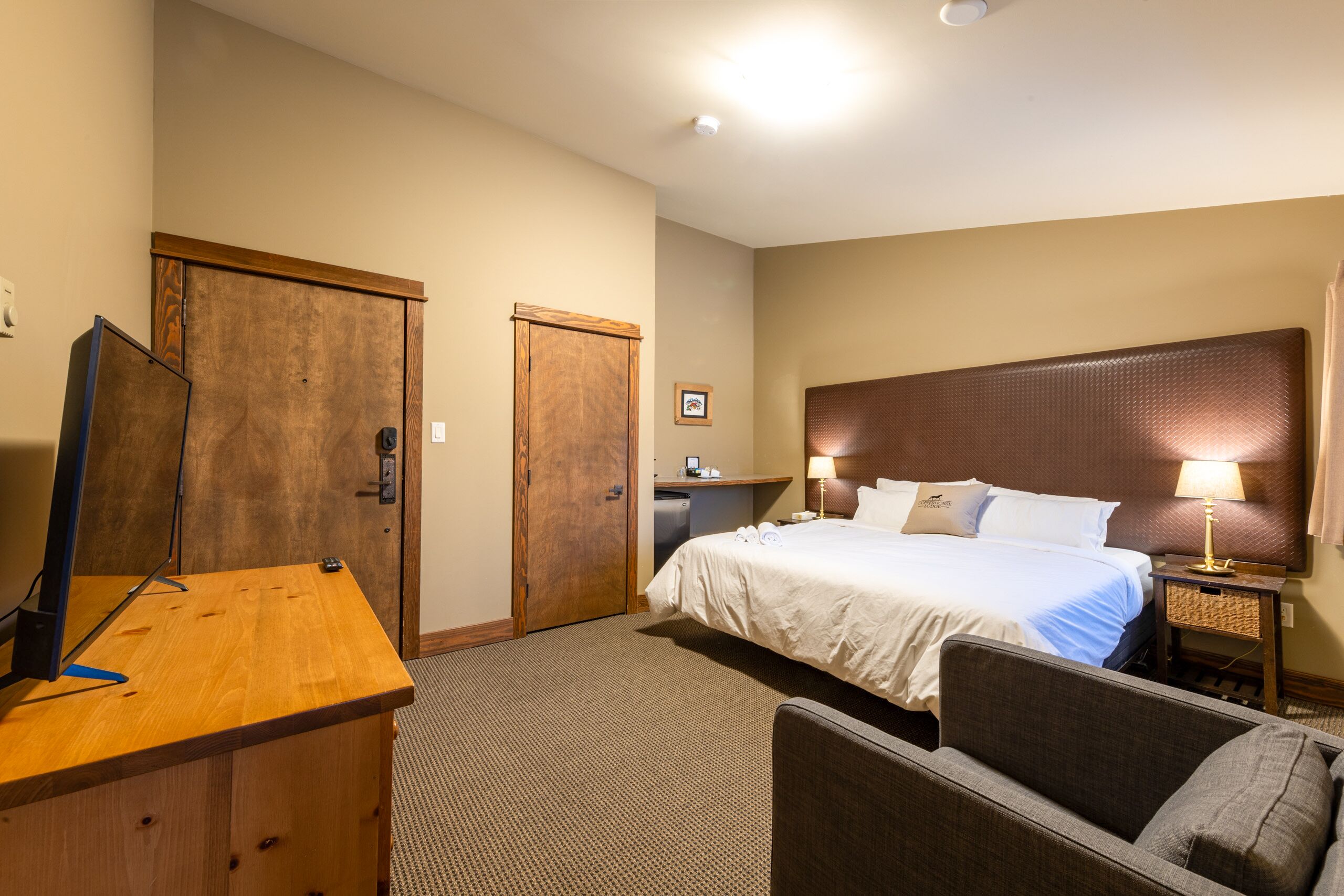 A hotel room with a bed, desk, chair, and a small table with a lamp.