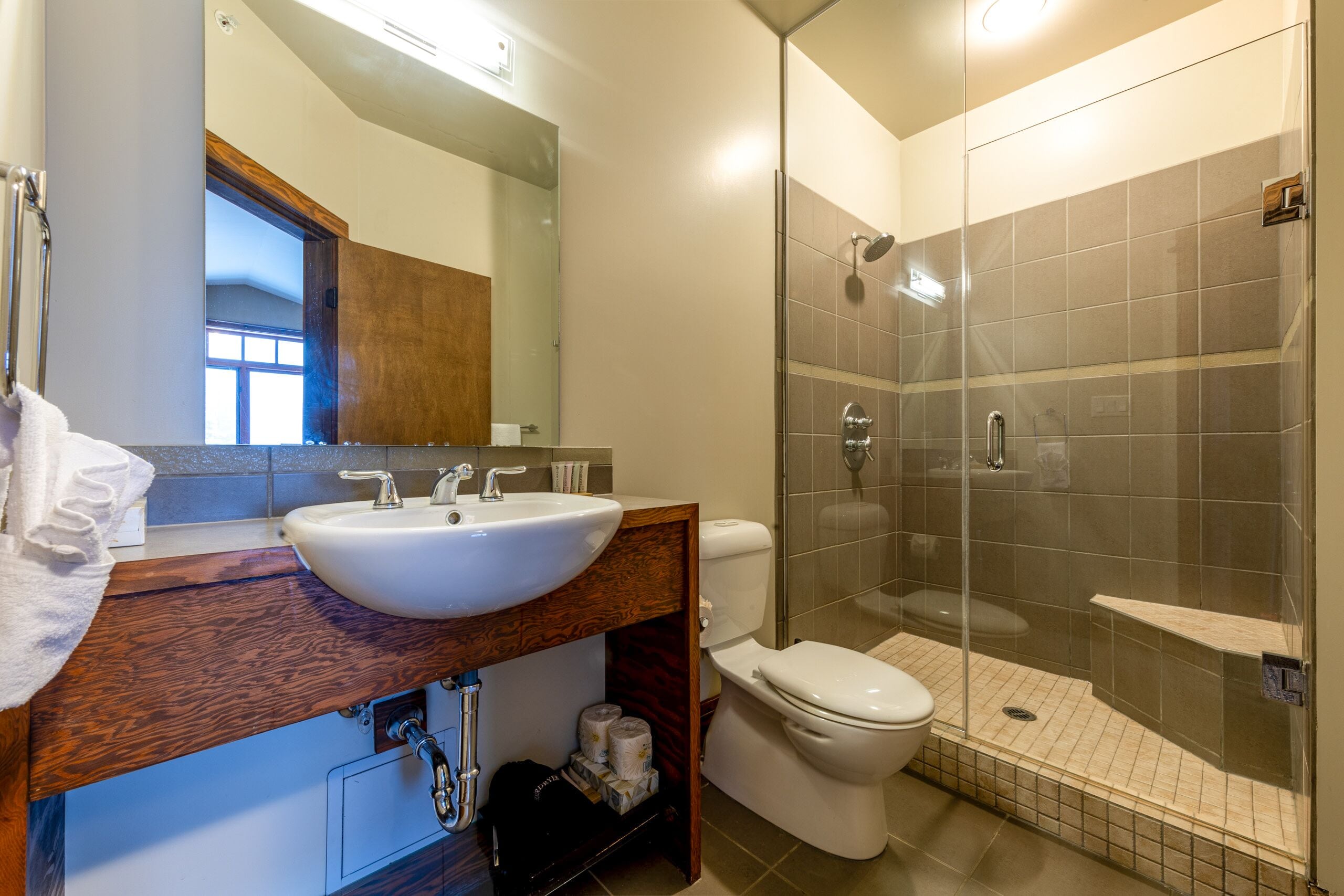 A modern bathroom with a wooden vanity, a white sink, a toilet, and a glass-enclosed shower area.