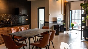 A modern kitchen with a stone backsplash, a black countertop, and a built-in stove. There is a dining area with a wooden table and leather chairs. The room has a large window with a view of buildings and a balcony with plants. - Reizz Residence by Plush (Kuala Lumpur)