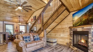 Un acogedor interior de cabaña con una escalera de madera, un sofá de cuero con cojines estampados, una chimenea y un cuadro con un paisaje.