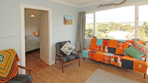A cozy living room with a wooden floor, a grey armchair, a colorful sofa, and a view through a large window. - Waiheke holiday home (Waiheke Island)