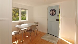 A dining area with a table and chairs, a dartboard on a door, and a window with a view of greenery. - Waiheke holiday home (Waiheke Island)