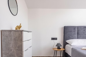 Un dormitorio minimalista con cama, cómoda, una mesita con lámpara y un enchufe en la pared.