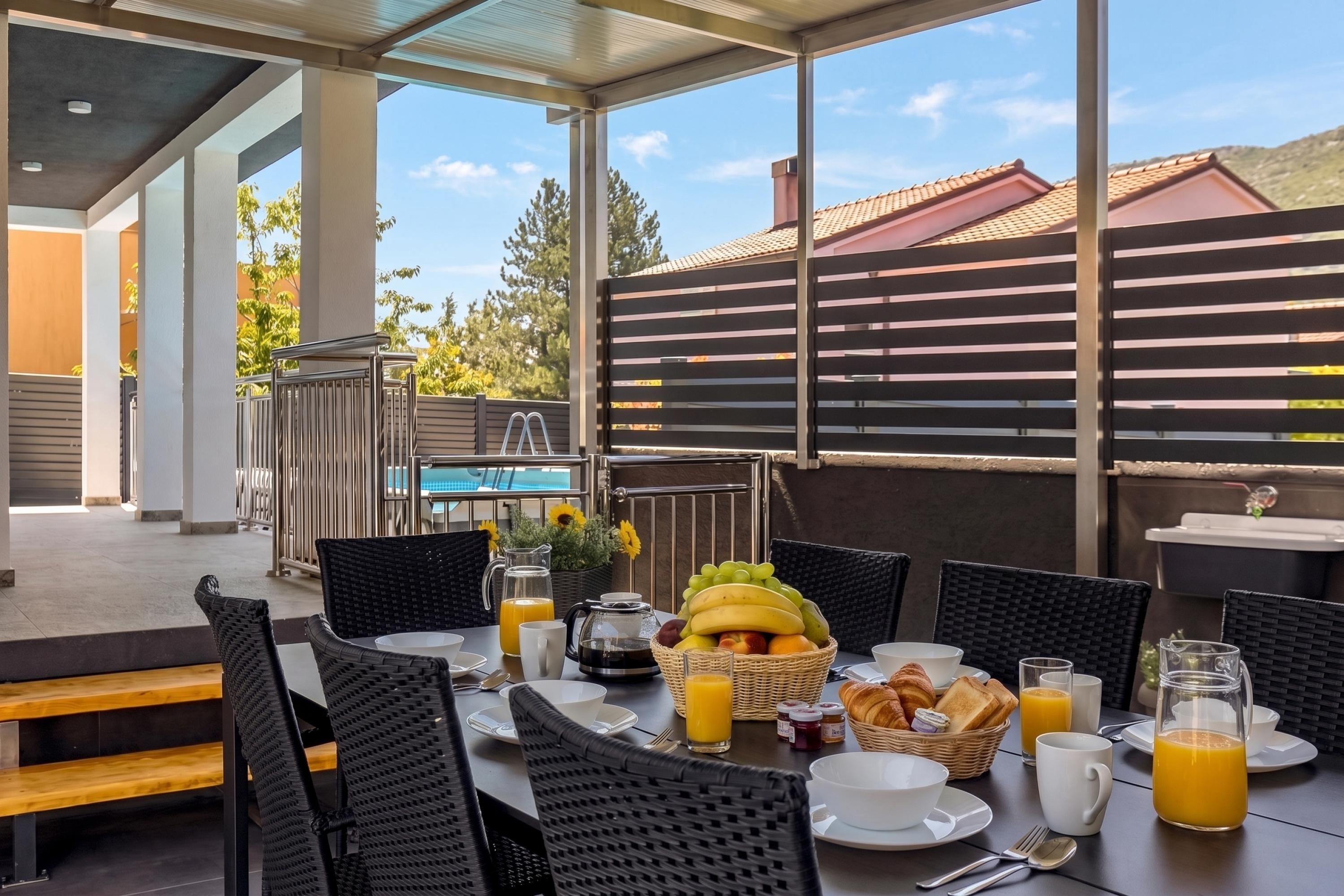 An outdoor dining area with wicker chairs and tables, a pool in the background, and a breakfast spread including fruits, bread, and beverages.